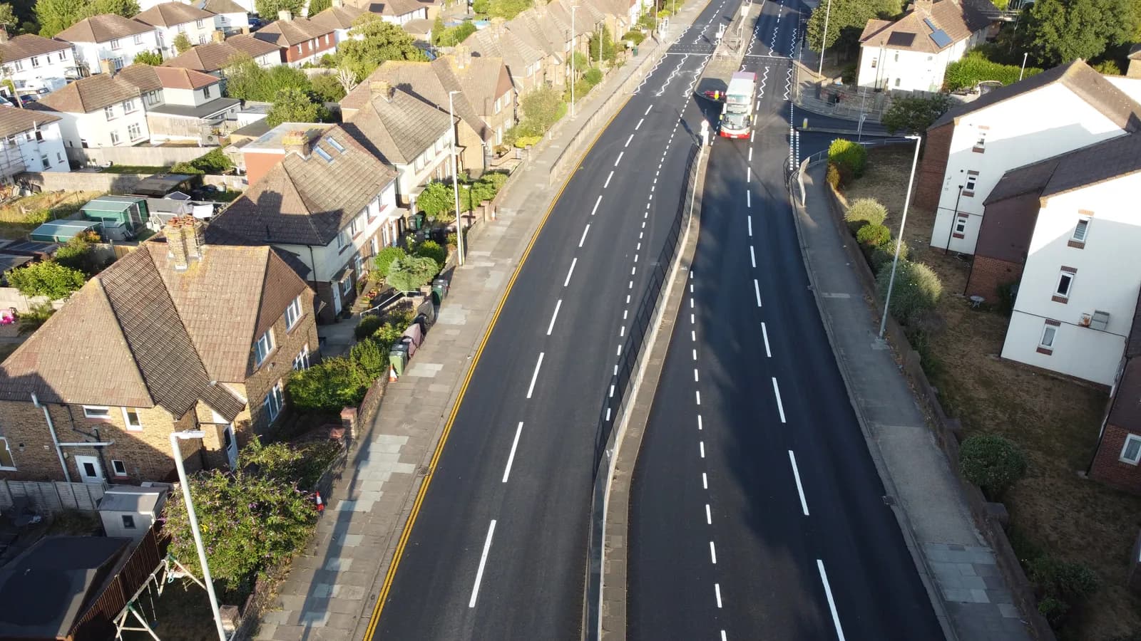 An aerial view shows a residential street with houses and trees lining a divided road, with a white truck visible on the right side.