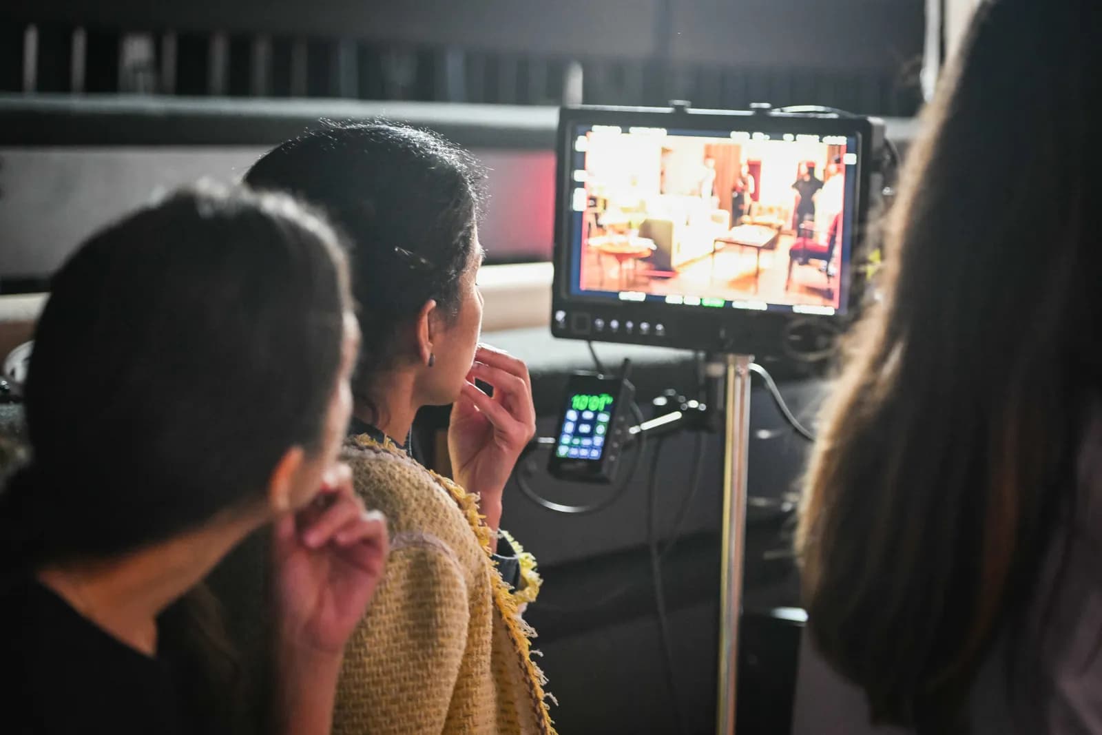Two people watch a monitor displaying an interior scene with furniture and a partial view of a person.