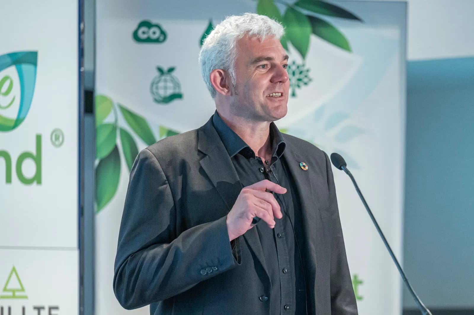 A man with gray hair in a dark suit speaks into a microphone at a podium, with green logos in the background.