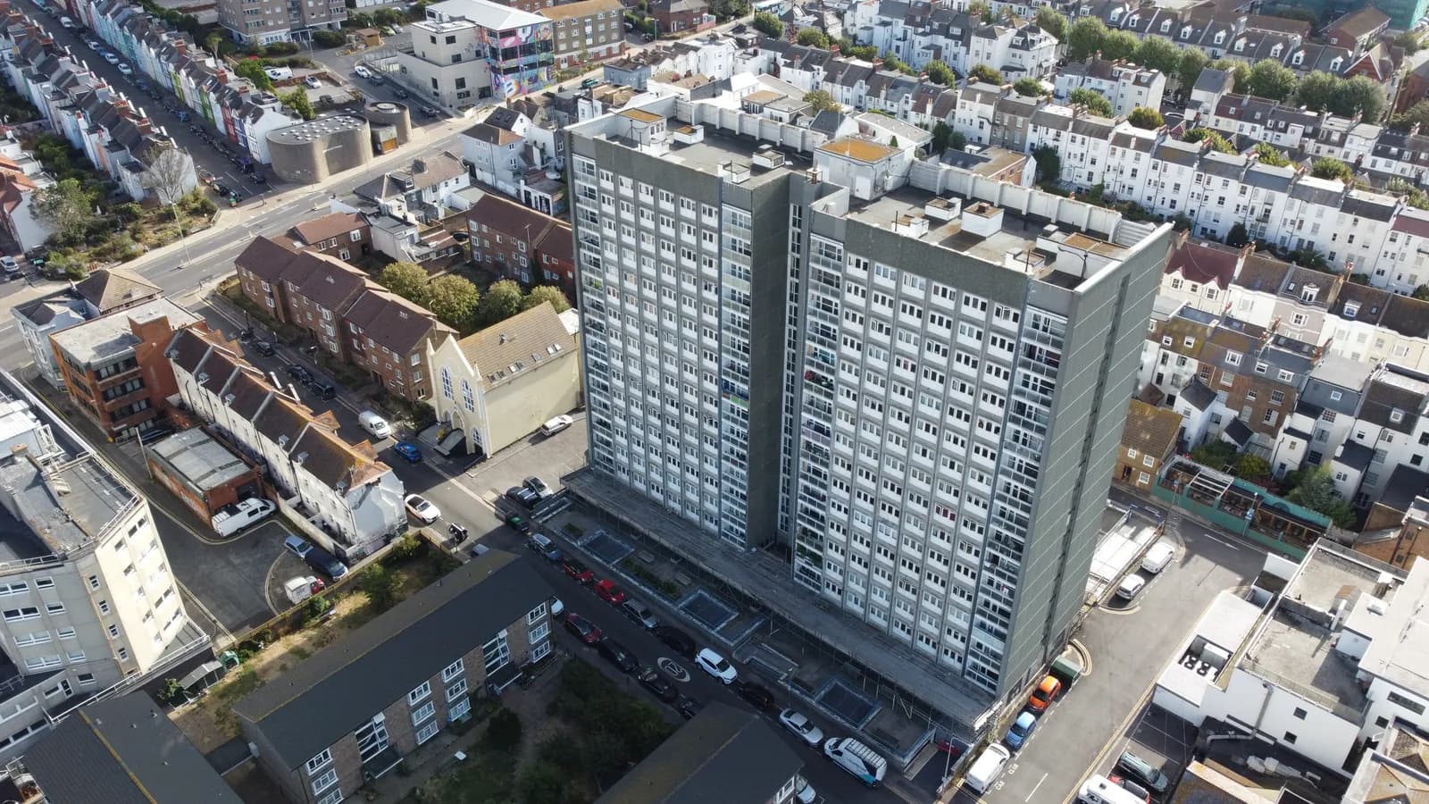 An aerial view of a large, gray high-rise apartment building surrounded by smaller residential buildings and streets with parked cars.